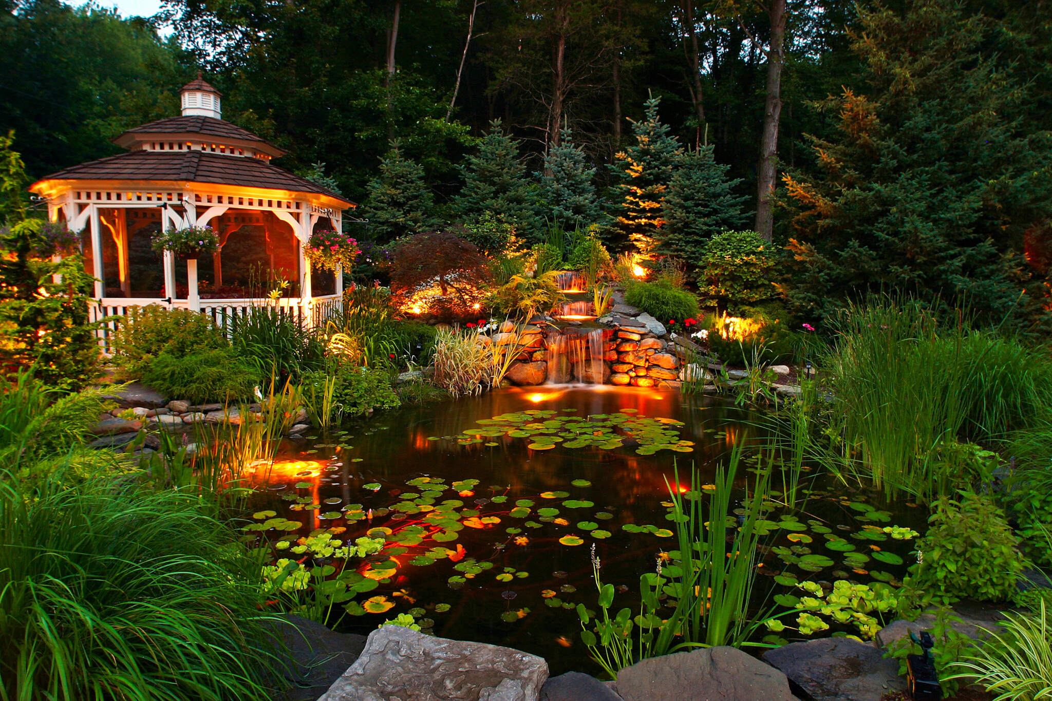 Luxury gazebo design with a charming gazebo, illuminated pond, cascading waterfall, and lush garden landscaping at night