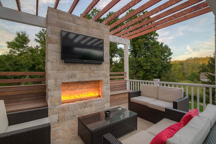 Luxury patio design with a cozy seating area, stone fireplace, mounted TV, and pergola, surrounded by lush greenery..jpg