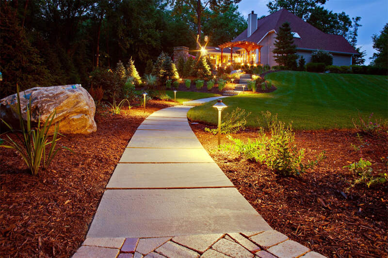 Illuminated stone walkway leading to a well-lit garden, showcasing elegant landscape design with premium hardscape elements