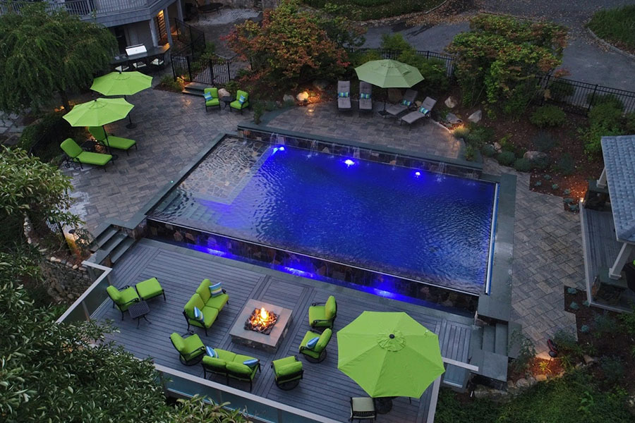 A stunning pool deck with green-cushioned seating, umbrellas, a fire pit, and an illuminated pool, surrounded by lush landscaping