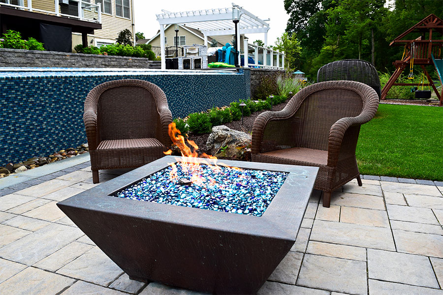 Connecting a pool with outdoor spaces featuring a fire pit, wicker chairs, and lush landscaping with a children's play area nearby