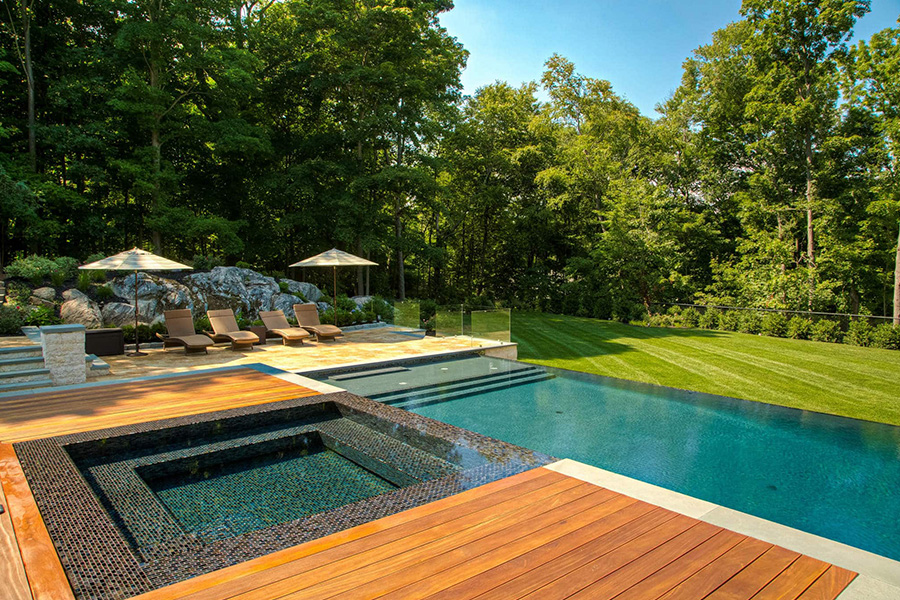 Luxury pool decking with wood and stone, surrounded by lounge chairs and lush greenery, creating a serene outdoor retreat.