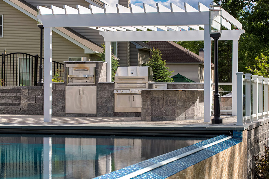 Outdoor space with a modern kitchen under a white pergola by the pool, featuring a grill, pizza oven, and stainless steel appliances.